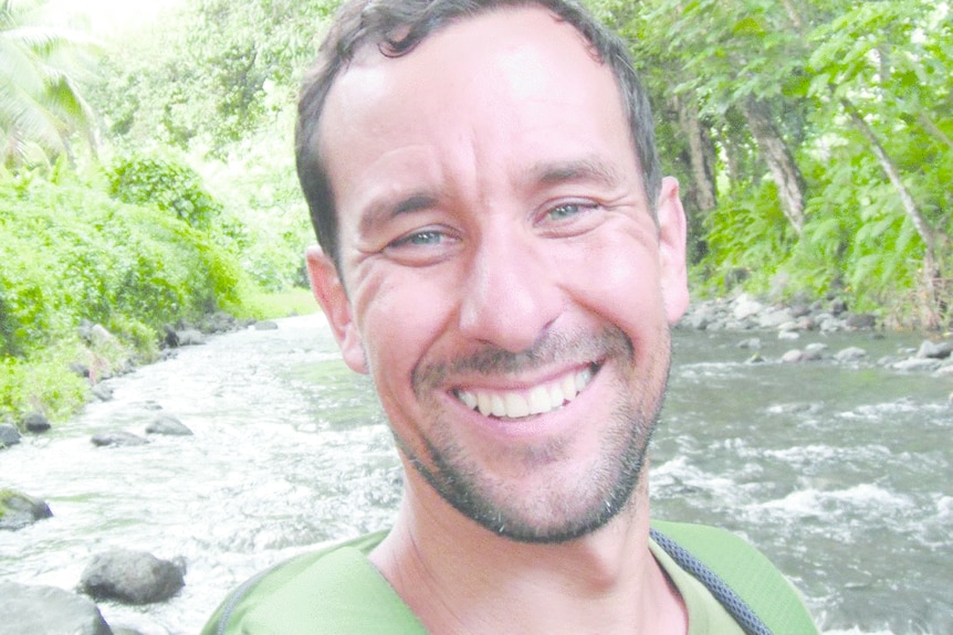 Close-up shot of Ross Frylick with short brown hair smiling widely, with river and bushland behind him.