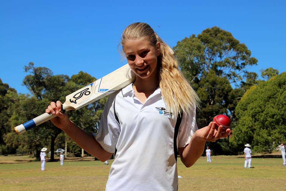 A girl holds a cricket ball in one hand and has a bat slung over her shoulder. A game of cricket is happening in the background.