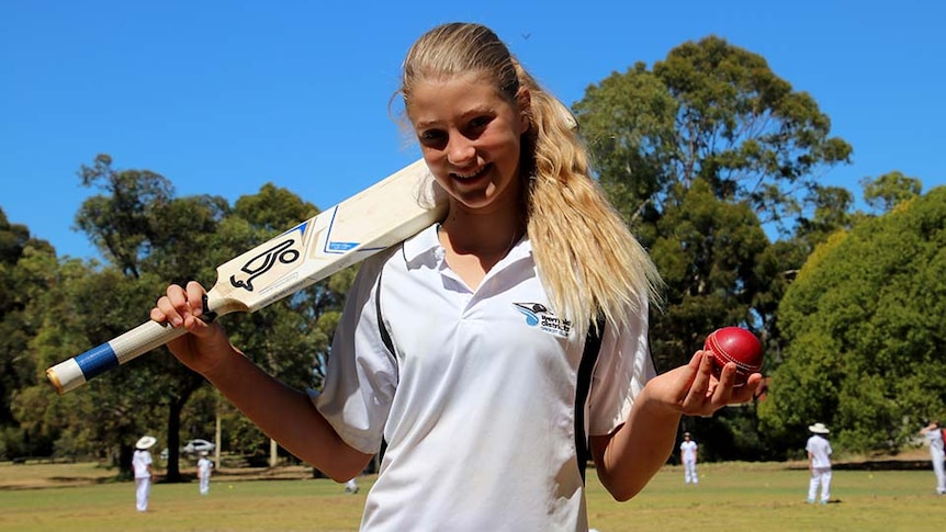 A girl holds a cricket ball in one hand and has a bat slung over her shoulder. A game of cricket is happening in the background.