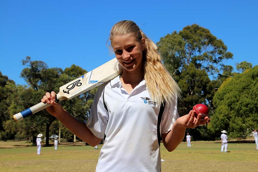 A girl holds a cricket ball in one hand and has a bat slung over her shoulder. A game of cricket is happening in the background.