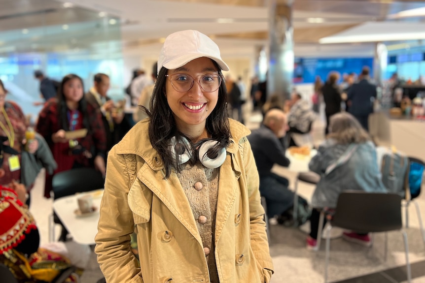 A woman wearing a cap, with headphones around her neck, in a crowded airport terminal.