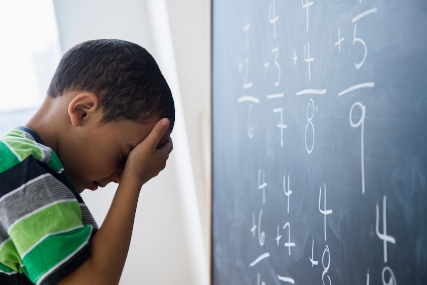 A boy holding his head in his hand in front of a blackboard.