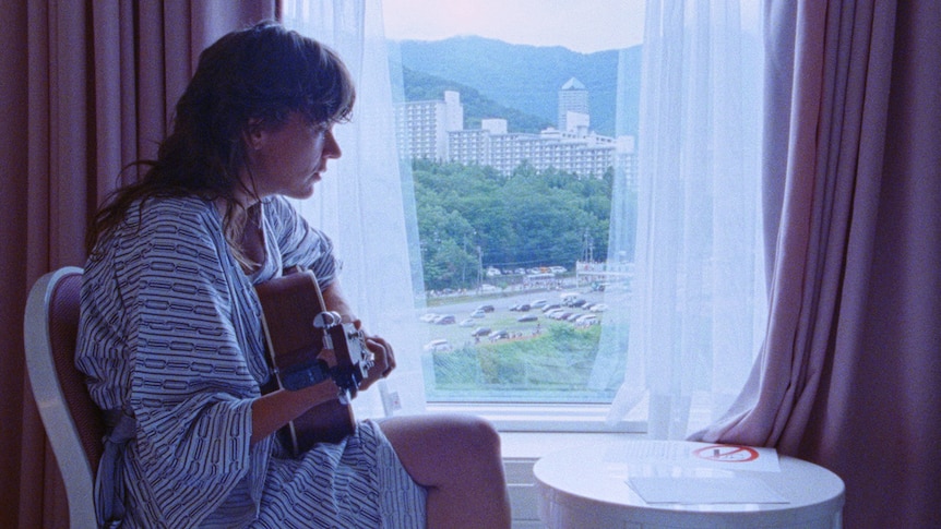 Courtney Barnett sits in a hotel room in a robe playing an acoustic guitar
