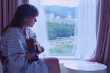 Courtney Barnett sits in a hotel room in a robe playing an acoustic guitar