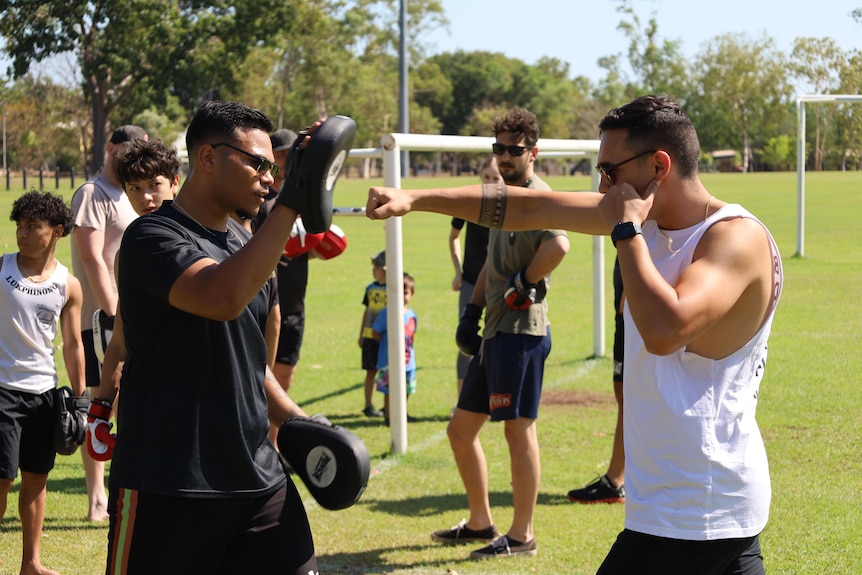Two men in sunglasses are pictured, one holds boxing pads while the other's arm is stretched out to hit the pad.