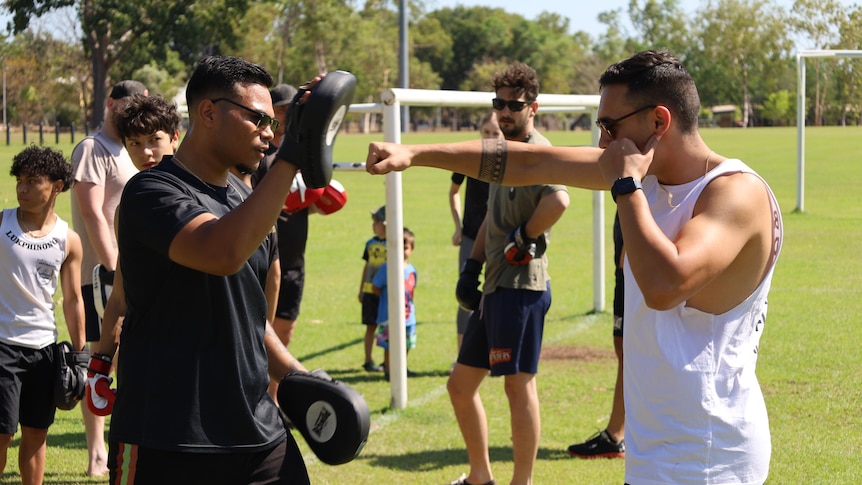 Two men in sunglasses are pictured, one holds boxing pads while the other's arm is stretched out to hit the pad.