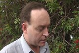 Head and shoulders shot of middle aged man with receding hairline standing near red flowering foliage.