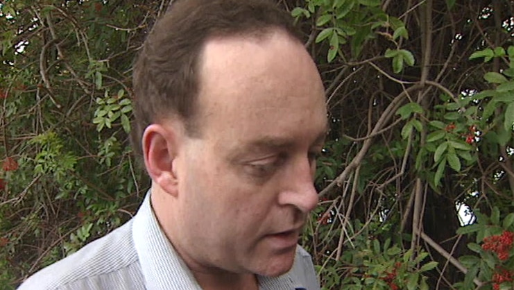 Head and shoulders shot of middle aged man with receding hairline standing near red flowering foliage.