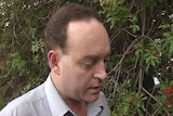 Head and shoulders shot of middle aged man with receding hairline standing near red flowering foliage.