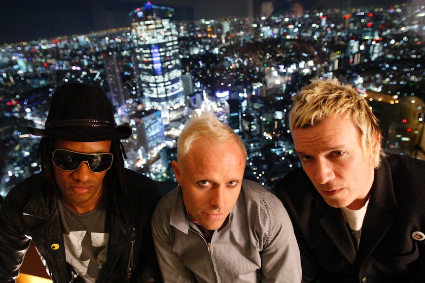 Maxim in sunglasses, Keith Flint and Liam Howlett in front of a glass window with the Tokyo night skyline in the background
