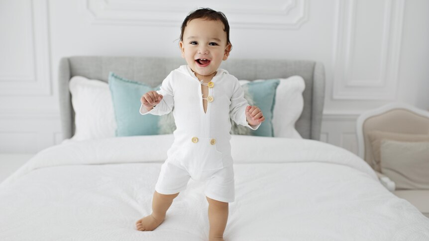 A smiling, dark-haired toddler standing on a bed.