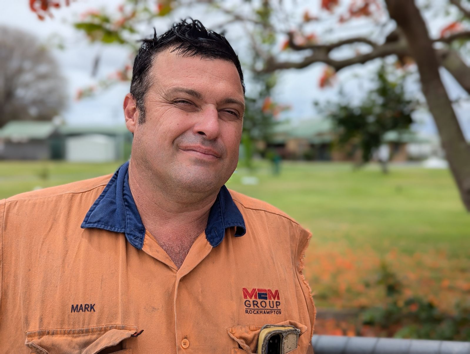 A man wearing a tradesman's shirt, squinting in the sun.