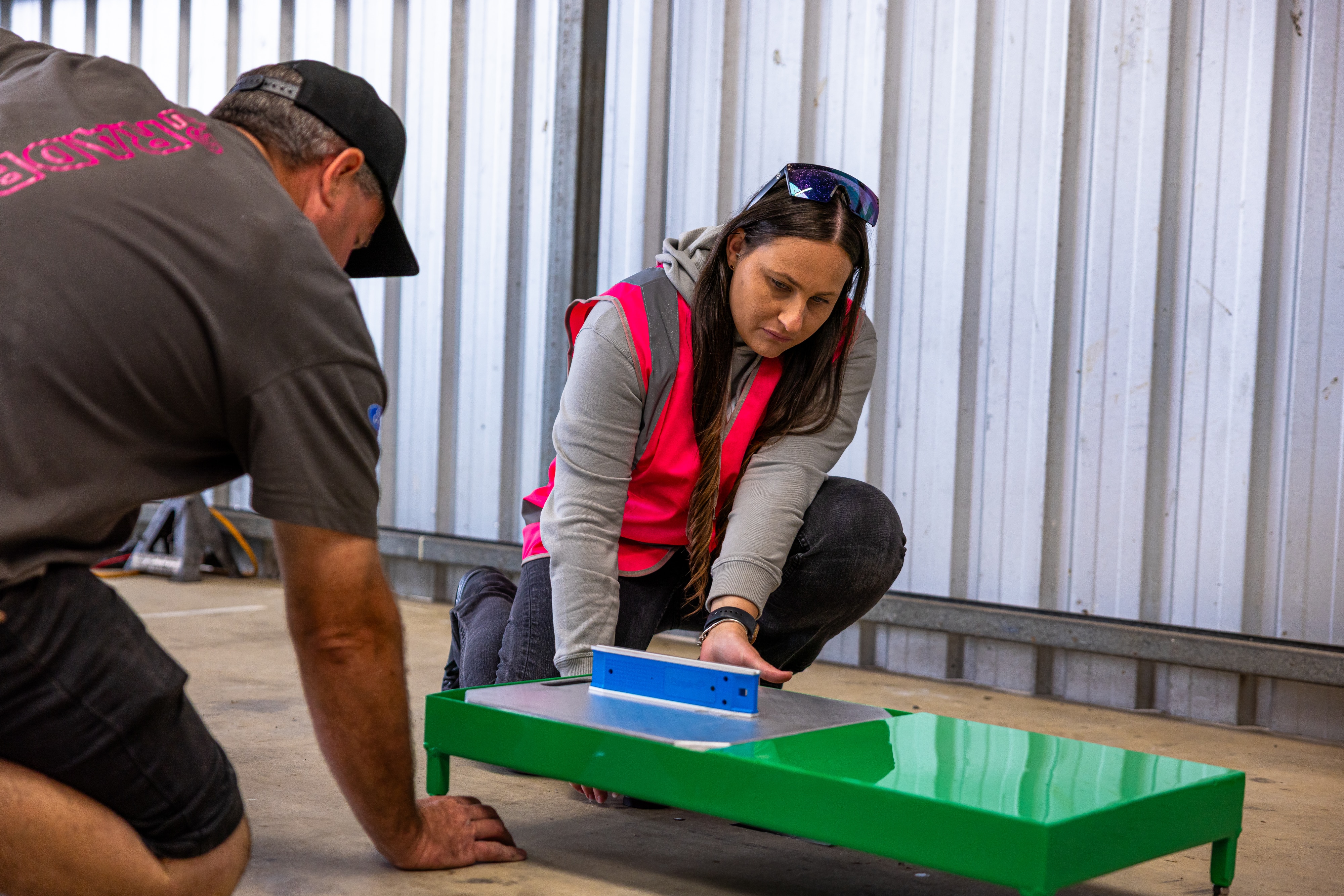 Natasha usa um colete rosa de alta visibilidade e está ajoelhada, trabalhando em uma peça verde do carro.