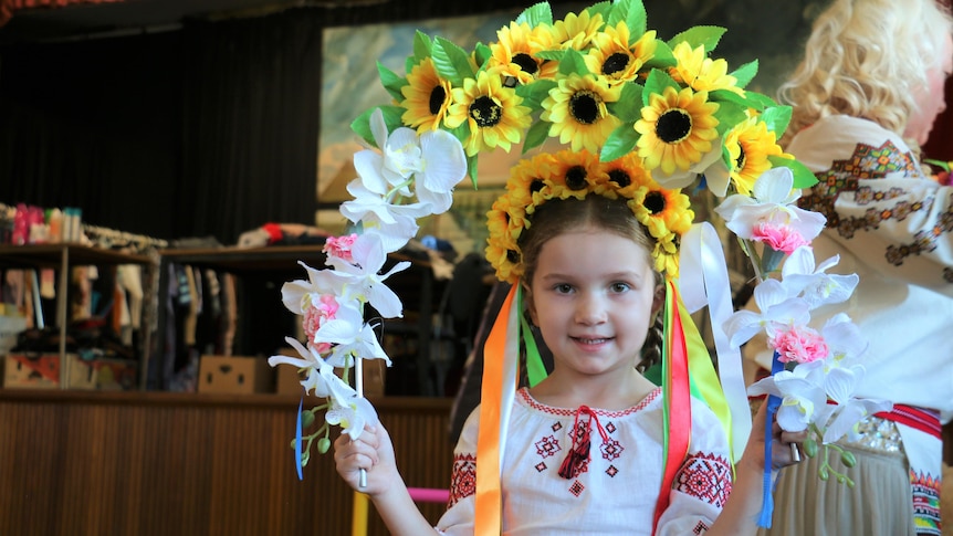 A young Ukrainian girl dressed in traditional dress and holding a an arch of flowers while smiling at the camera.