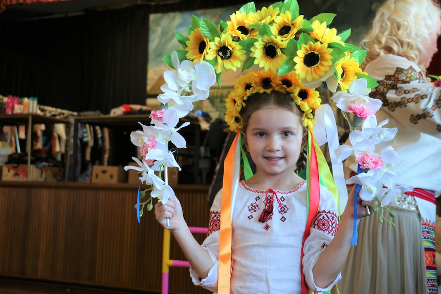 A young Ukrainian girl dressed in traditional dress and holding a an arch of flowers while smiling at the camera.