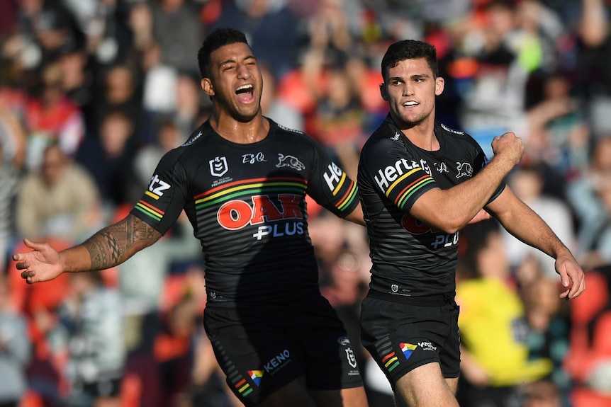 A male NRL player has his right arm across his body as he celebrates a try, while a teammate approaches him to congratulate him.