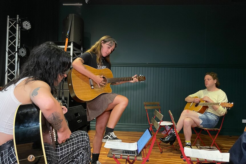 A woman plays the guitar with one foot on a chair as students watch on. 