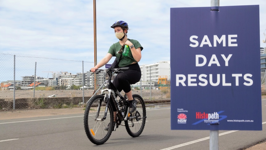 A woman wears a mask while riding a bike past a COVID testing clinic sign that reads "same day results"