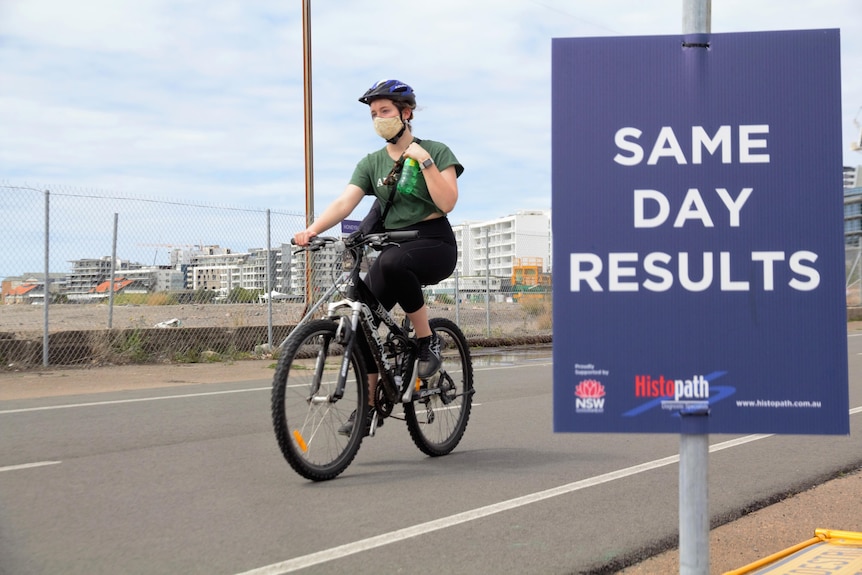 A woman wears a mask while riding a bike past a COVID testing clinic sign that reads "same day results"