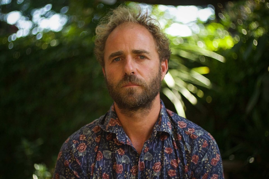 A man with a beard looks into the camera with a serious expression on his face. A garden can be seen in the background.