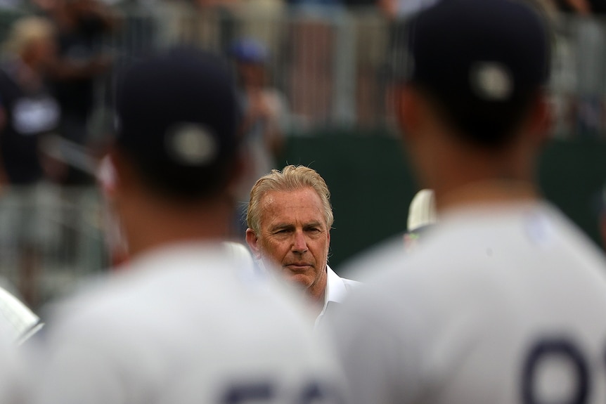 Kevin Costner is seen between two players, blurred in the foreground