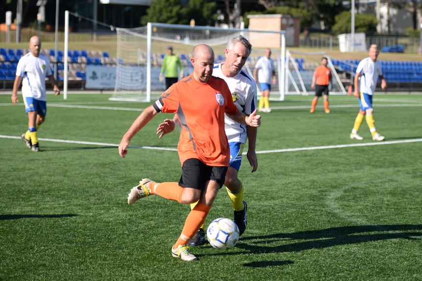 A man in an orange shirt playing football, he is about to kick the soccer ball and a man in a white shirt is behind him