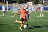 A man in an orange shirt playing football, he is about to kick the soccer ball and a man in a white shirt is behind him