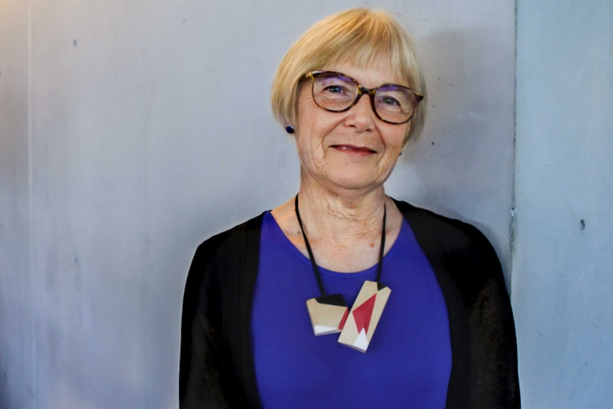Older woman with glasses and blonde hair standing in front of grey wall.
