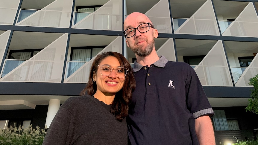 A man and a woman stand in front of a new apartment block and smile.