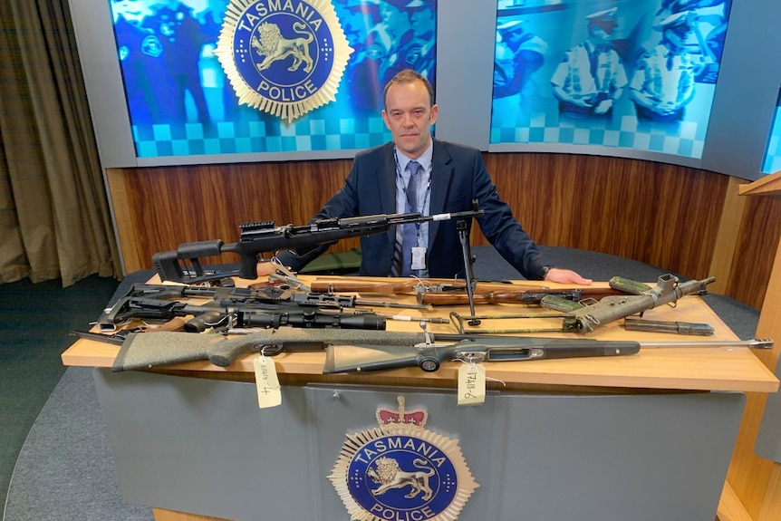 A police officer stands behind a table covered in guns.