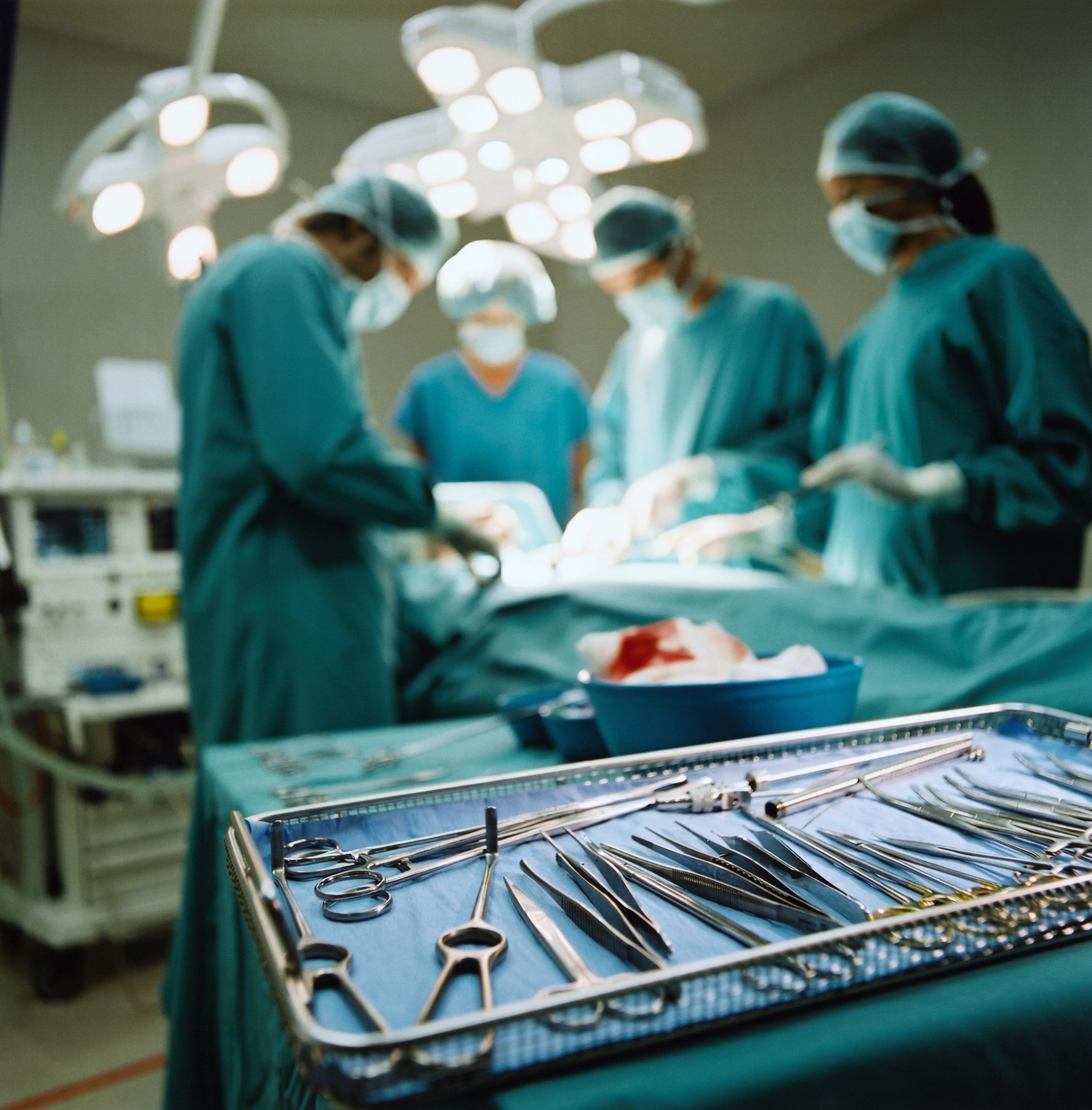 In the foreground, a tray of medical instruments, in the background four surgeons wearing scrubs operate on a patient.