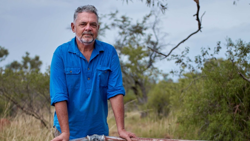 A man in a blue shirt stands in the NT outback, leaning on a decorate, horizontal metal pole