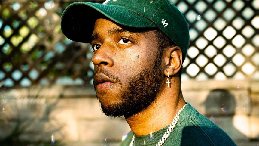 Portrait photo of 6lack looking to the left, wearing a green cap, green t-shirt, and diamond chain.