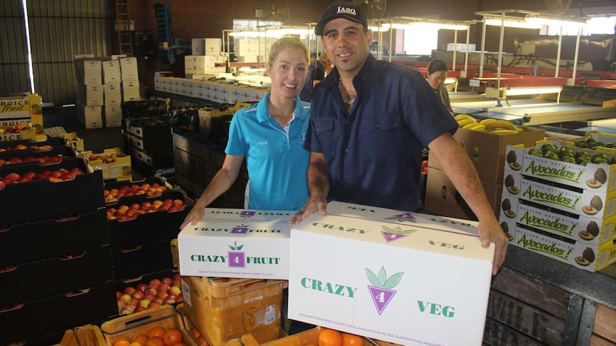 A man and a women stand with amid boxes of fruit and vegetables.