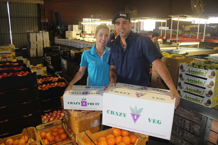 A man and a women stand with amid boxes of fruit and vegetables.