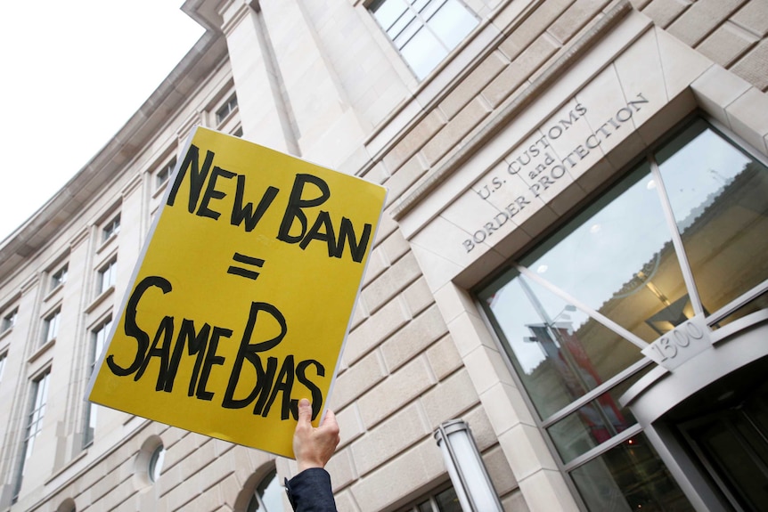 Protesters hold a sign reading "new ban = same bias"