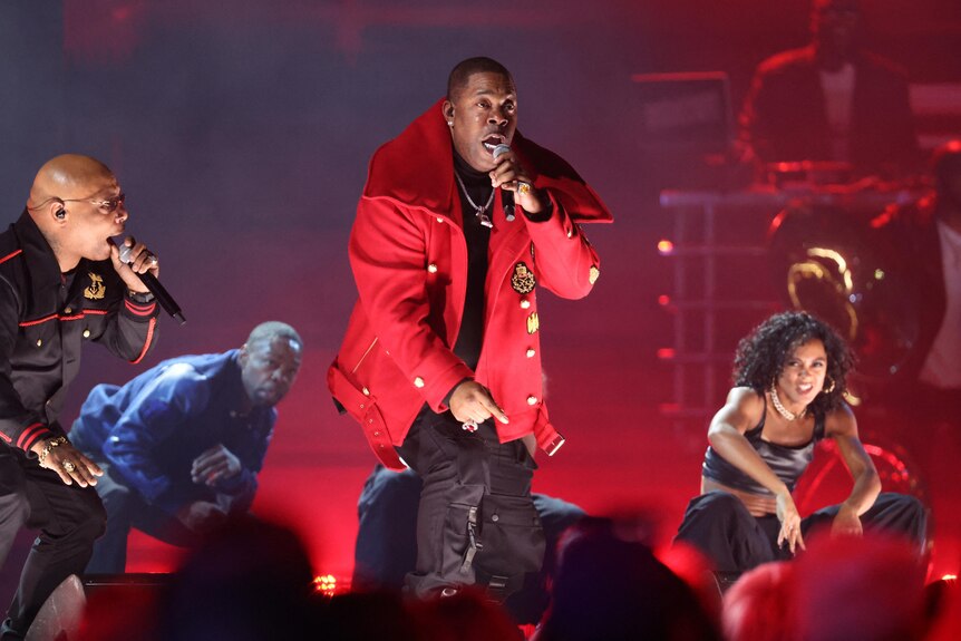 Busta Rhymes on stage rapping in a red coat with people surrounding him on stage
