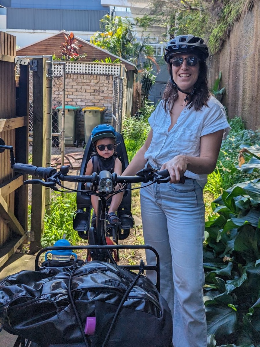 A baby in a child seat on a bike with a woman holding the handlebars wearing a helmet. 