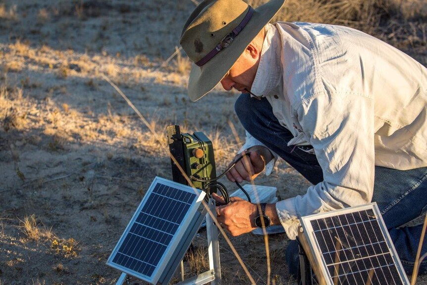 A man wearing a broad-brimmed hat crouches in the afternoon light to work on an electrical device fitted with solar panels.