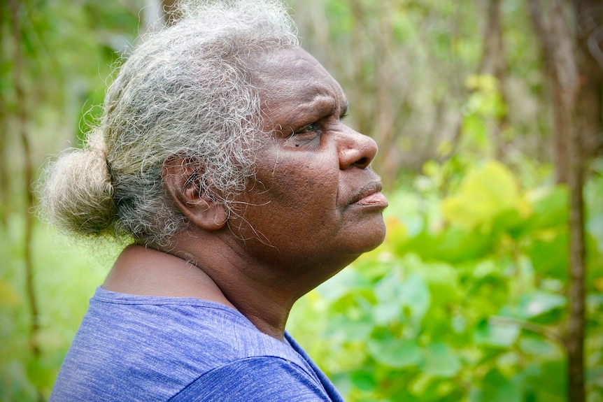 An Aboriginal woman in a purple shirt looking up, with greenery in the background. 