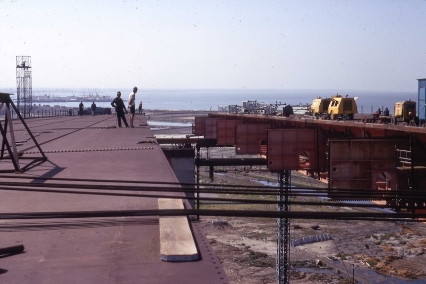 Workers stand on a large metal platform, port in background.