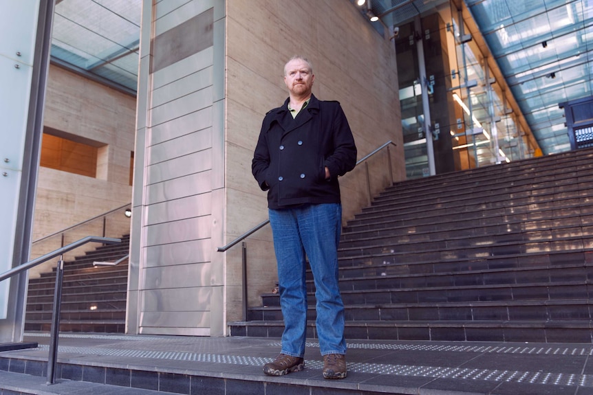 A wide shot portrait of a man standing on the stairs in a nondescript urban background.