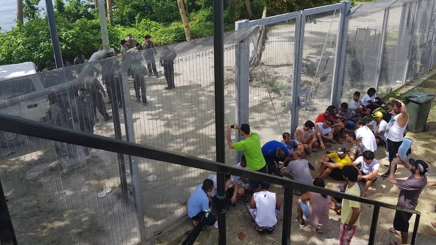 Refugees and asylum seekers are sitting on one side of a gate while guards are standing on the other side.