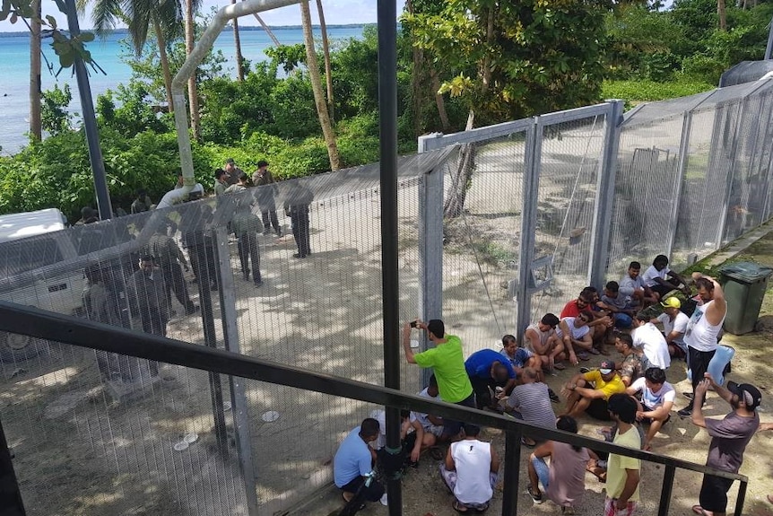 Refugees and asylum seekers are sitting on one side of the gate while guards are standing on the other side.