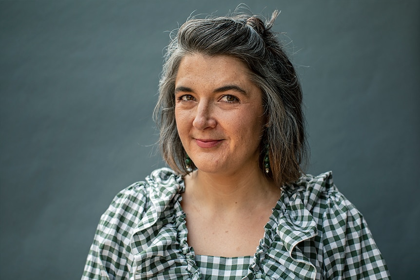 A woman with short dark and grey hair in white and dark green checked shirt stands smiling in front of grey background.