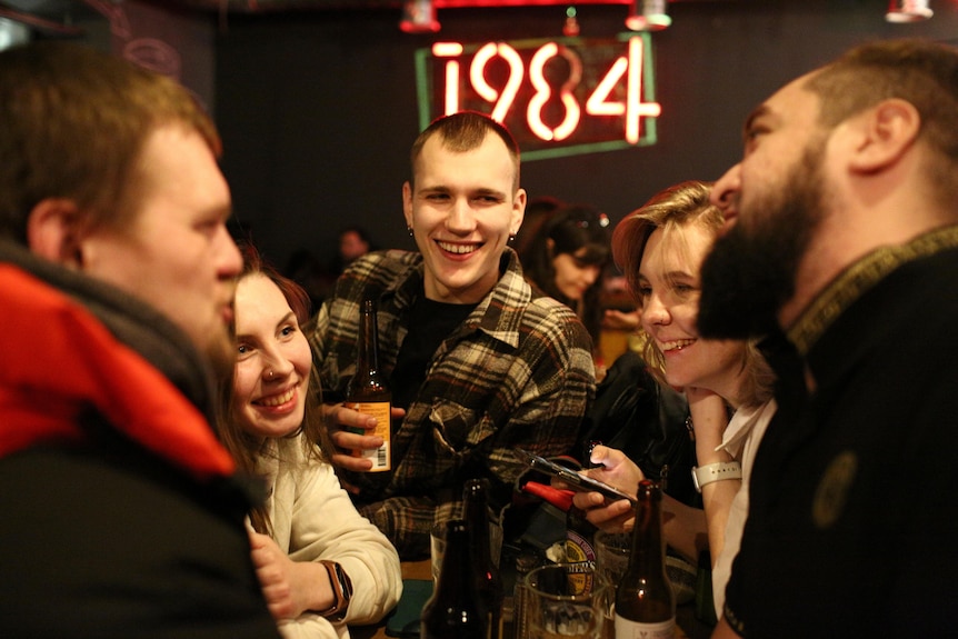 A group of five young people are standing in a circle relaxing and laughing in a dimly-lit bar.