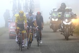 Three top riders from the Tour de France race through the mist on Col du Portet, with headlights from motorbikes in background.