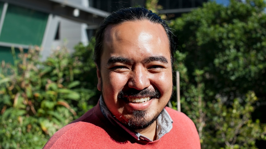 Adam Liaw, smiling widely, wears peach coloured jumped and stands outside with green bushes in the background.