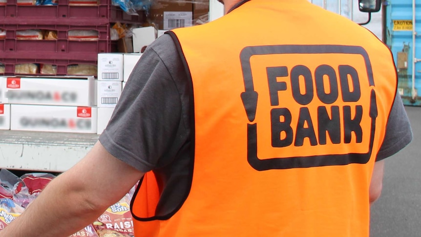 A Foodbank worker wearing an orange vest with insignia on back carrying a crate of Mighty Soft bread.