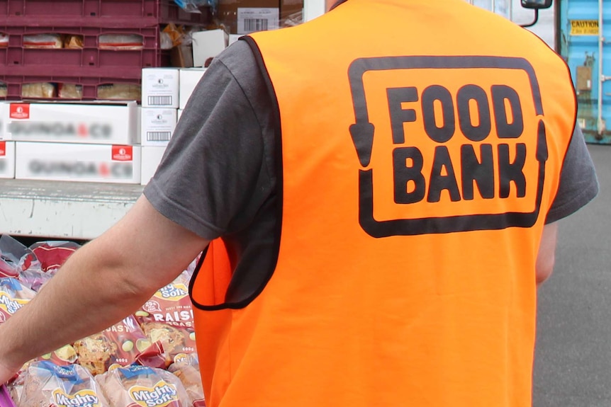 A Foodbank worker wearing an orange vest with insignia on back carrying a crate of Mighty Soft bread.